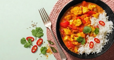 Groene curry met rijst, geserveerd in een zwarte kom, met kleurrijke groenten zoals paprika, bloemkool en tofu, en gegarneerd met koriander en rode peper, met een vork en kruiden ernaast op een roze onderzetter.