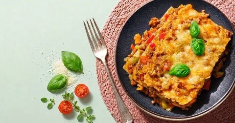 Lasagne op een zwart bord, gegarneerd met verse basilicum. Naast het bord liggen een vork, geraspte kaas, basilicumbladeren, cherrytomaatjes en oregano op een lichtgroene ondergrond.
