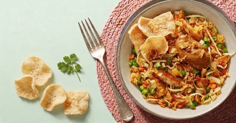 Bord met nasi goreng, kip, kroepoek, satésaus en groenten, geserveerd op een roze placemat met kroepoek en koriander als garnering.