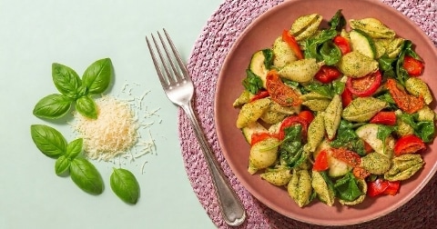 Pastasalade met pesto, spinazie, tomaat, courgette en basilicum, geserveerd op een roze bord met een vork erbij. Daarnaast liggen verse basilicumblaadjes en geraspte kaas.