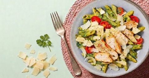 Pasta met pesto, gegrilde kip, cherrytomaatjes en Parmezaanse kaas op een bord, met een vork en extra kaas en peterselie ernaast.