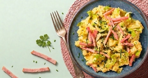Stamppot met aardappel, gesneden hamreepjes, andijvie en bieslook op een blauw bord, met een metalen vork ernaast.