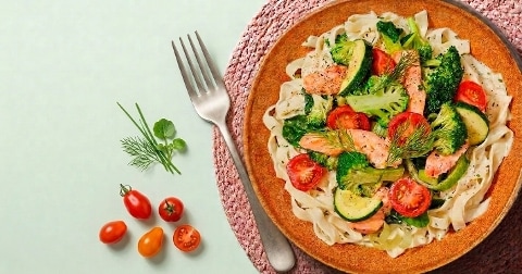 Tagliatelle met broccoli, courgette, cherrytomaatjes en stukjes zalm, geserveerd op een bord met een vork ernaast en verse kruiden en tomaatjes op tafel.