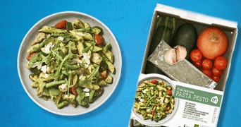 Italiaanse pasta pesto maaltijdpakket van Albert Heijn met verse ingrediënten zoals pasta, tomaten, courgette, avocado, en een zakje pesto, afgebeeld naast een bereid gerecht van pasta pesto.
