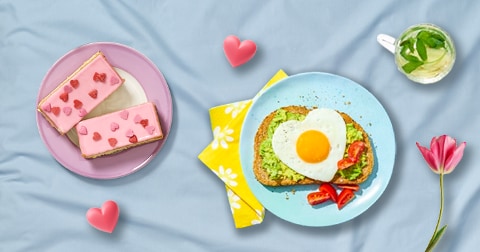 Ontbijtgerecht met een boterham belegd met avocado en een hartvormig gebakken ei, daarnaast roze tompoucen met hartjes en een glas met een frisse drank en muntblaadjes, op een dienblad gepresenteerd op een dekbed.