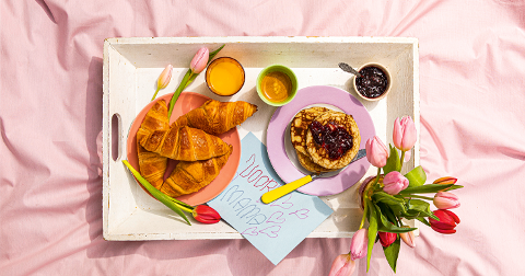 Ontbijt op bed met croissants, pannenkoeken, sinaasappelsap, tulpen en een kaartje met "Voor mama".