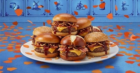 Een bord met acht cheeseburgers met bacon en gefrituurde uitjes, geserveerd op een blauwe ondergrond met oranje confetti, met tegels met Hollandse motieven op de achtergrond.