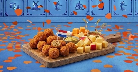 Versierde borrelplank met bitterballen, kaasblokjes, mosterd en een Nederlands vlaggetje, omringd door oranje confetti, tegen een achtergrond van blauw Hollandse tegels.