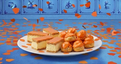 Schotel met oranje tompoucen en soesjes op een blauwe tafel, versierd met oranje confetti, typisch voor Koningsdag, met Nederlandse tegels op de achtergrond.