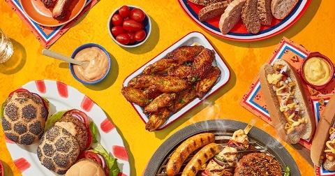 Een kleurrijk tafelbeeld met verschillende barbecuegerechten, waaronder hamburgers, kippenvleugels, worstjes, spiesjes, een hotdog met saus, brood, cherrytomaten en een bakje dipsaus.
