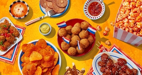 Een feestelijk assortiment Nederlandse snacks en hapjes op een gele achtergrond, waaronder bitterballen met Nederlandse vlaggetjes, chips, popcorn, gehaktballetjes, kipstukjes en plakjes worst, geserveerd met verschillende dips.
