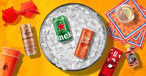 Blikje Heineken en Viva Tinto in een schaal met ijs, omringd door feestelijke accessoires zoals rode zonnebril, bekers, servetten met een glas citroenwater, en blikjes Coca-Cola en Lavish Passiontin, op een gele achtergrond.