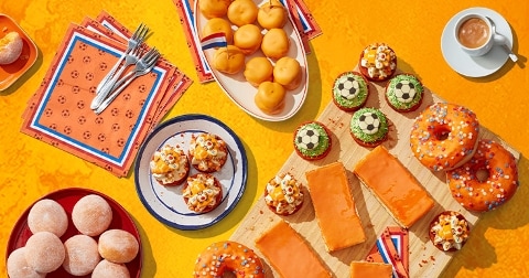 Feestelijke oranje gebakjes, donuts en tompoucen op een tafel, aangevuld met voetbaldecoraties en servetten, perfect voor Koningsdag of een EK voetbalfeest.