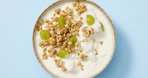 Kommetje yoghurt met granola, groene druiven en hazelnoten op een blauwe ondergrond.
