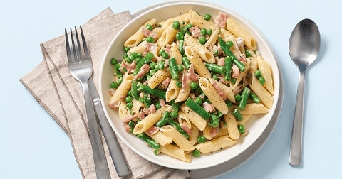 Penne pasta met doperwten, sperziebonen en stukjes ham op een wit bord, geserveerd met een vork en lepel op een lichtblauwe achtergrond.