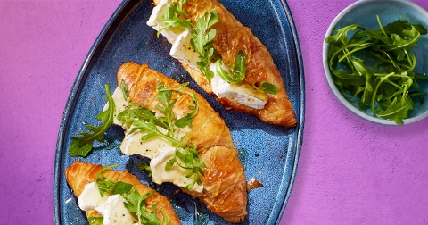 Drie croissants belegd met brie en rucola op een blauw bord, met een schaaltje rucola ernaast op een paarse achtergrond.