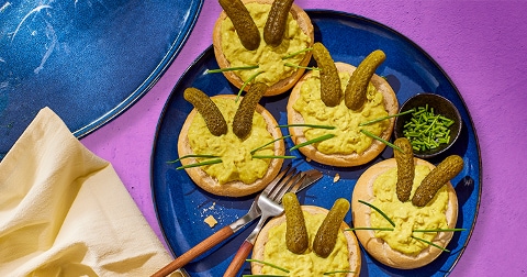 Rond broodbeleg op een blauw bord, versierd als konijnengezichtjes met smeuïge spread, augurken als oren en bieslook als snorharen, geplaatst op een paarse achtergrond.
