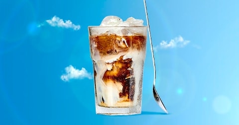 IJskoffie met melk in een glas met ijsblokjes tegen een blauwe achtergrond met wolken, en een lange lepel ernaast.