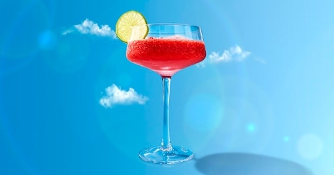 Een glas met een rode cocktail of slush, gegarneerd met een schijfje limoen, tegen een blauwe lucht met enkele wolken.