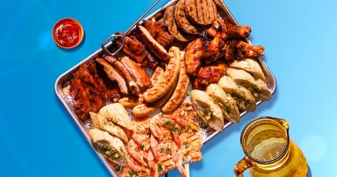 Grote schaal met gegrild vlees, waaronder worstjes, spareribs, kippenvleugels, hamburgers, garnalenspiesen en stukken kruidenstokbrood, geserveerd met een bakje ketchup en een kan bier op een blauwe achtergrond.