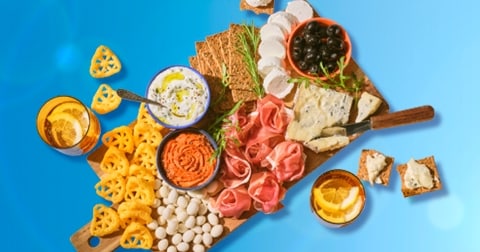 Borrelplank met diverse hapjes zoals kazen, olijven, crackers, dipjes, vleeswaren, snacks en drankjes met citroen, gepresenteerd op een houten plank tegen een blauwe achtergrond.