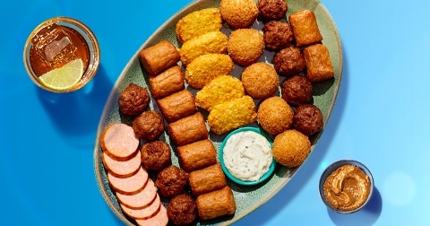 Schotel met diverse borrelhapjes, waaronder gesneden worst, bitterballen, mini saucijzenbroodjes, kaassoufflés, dip en pindasaus, geserveerd met een glas frisdrank met citroen op een blauwe achtergrond.