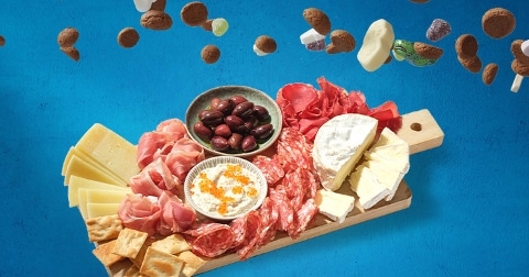 Borrelplank met onder andere kaas, vleeswaren, olijven en crackers op een blauwe achtergrond, met vallende pepernoten en snoepjes erboven