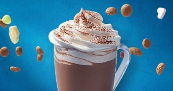 Glas warme chocolademelk met slagroom en cacaopoeder, omringd door kruidnoten, schuimpjes en stukjes witte chocolade tegen blauwe achtergrond