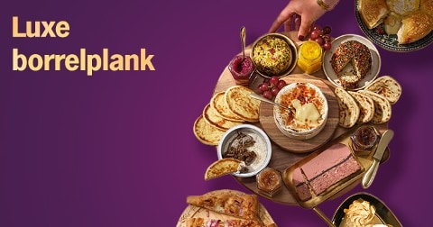 Luxe borrelplank met diverse toastjes, dips, paté, kaas en brood op een houten plank tegen een paarse achtergrond, met een hand die iets pakt