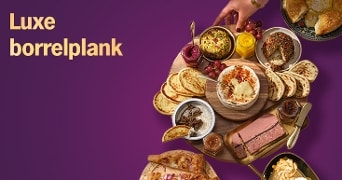 Luxe borrelplank met diverse toastjes, dips, paté, kaas en brood op een houten plank tegen een paarse achtergrond, met een hand die iets pakt