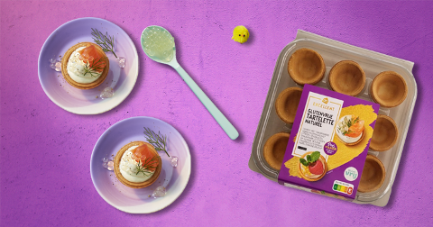 Twee gevulde glutenvrije tartelette taartjes met zalm, dille en roomkaas, op borden gepresenteerd naast een verpakking Albert Heijn Excellent glutenvrije tartelette naturel op een paarse ondergrond.