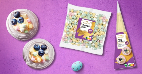 Twee meringue nestjes met blauwe bessen en gekleurde meringue drops, samen met een verpakking AH Excellent Meringue Drops en een spuitzak AH Excellent Romige Mousse op een paarse ondergrond.