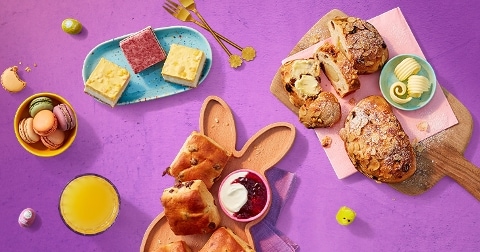 Paasontbijt met zoete broodjes, macarons, boter, jam, vruchtensap en gekleurde paaseieren op een paarse achtergrond.