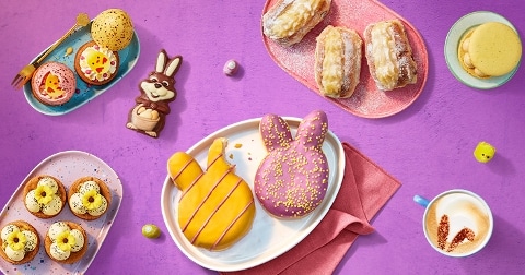 Paasgebakjes en zoetigheden op een paarse ondergrond, waaronder konijnvormige donuts, een chocolade paashaas, gevulde eieren, koffiebeker met latte art en diverse andere gebakjes.