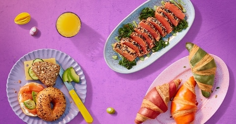 Ontbijt met bagel met zalm en avocado, meergranenbrood met kaas, plakjes zalm met sesam en zeewier, kleurrijke croissants en een glas sinaasappelsap, gepresenteerd op een paarse achtergrond.