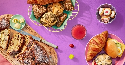 Ontbijt met diverse broodjes, croissant, zoet brood, boter, een glas sap en luxe petitfours op een paarse ondergrond.