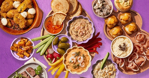 Een kleurrijke borrelplank met verschillende dips, groentes, mini broodjes, olijven, blokjes kaas, prosciutto, warme hapjes en spreads, gepresenteerd op een paarse ondergrond.