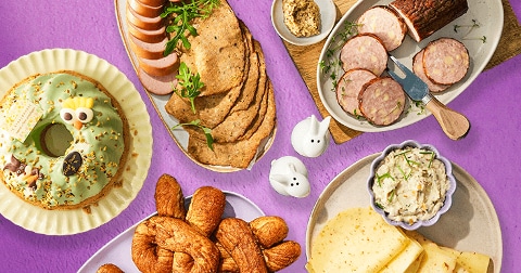 Verschillende paasproducten op een tafel, waaronder brood met groene glazuur en decoratie, plakken kaas, croissants, crackers en plakjes worst, geschikt voor een feestelijk ontbijt of brunch.
