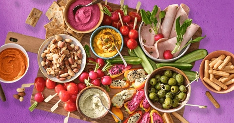 Borrelplank met groentedips, crackers, olijven, noten, radijsjes, groentespreads en plakjes vleeswaren op een paarse ondergrond.