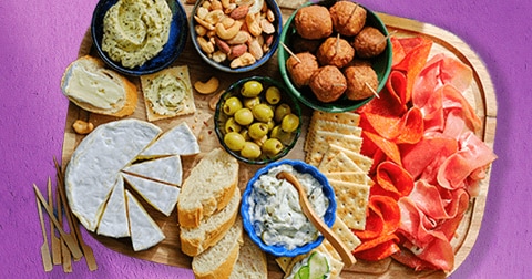 Borrelplank met brie, brood, olijven, crackers, noten, dip, kaas, falafel, en verschillende soorten vleeswaren op een houten plank tegen een paarse achtergrond.