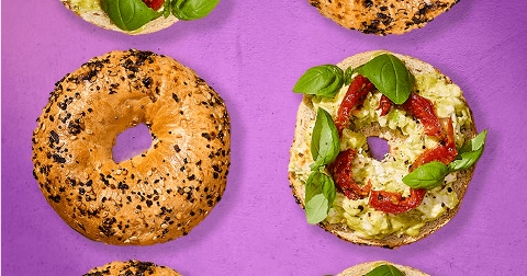 Bagels met sesamzaad en maanzaad, sommige belegd met avocadospread, zongedroogde tomaat en verse basilicum, op een paarse achtergrond.