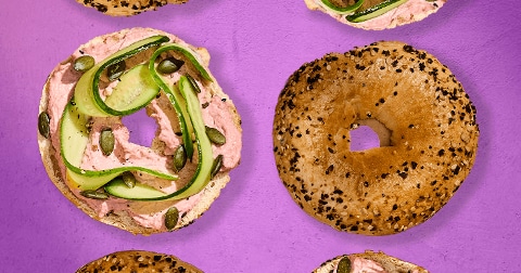 Bagel met roze smeersel, plakjes komkommer en pompoenpitten op een paarse achtergrond, naast een bagel met sesamzaad.