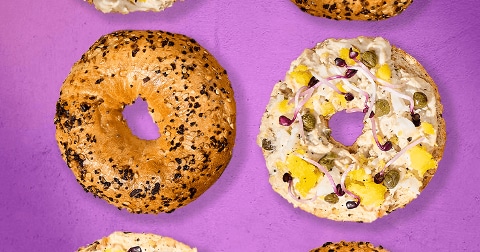 Bagels, waarvan één belegd met salade, kappertjes, ui en stukjes gele groente op een paarse achtergrond.