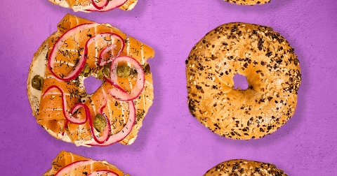 Bagels, waarvan één belegd met zalm, rode ui, kappertjes en kruiden, op een paarse achtergrond