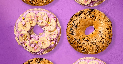 Bagels met sesamzaad en maanzaad, deels belegd met roze crème, plakjes banaan en vrolijke sprinkels, op een paarse ondergrond.