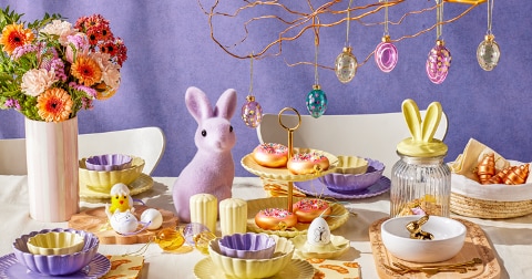 Paastafel vol met kleurrijke borden, kommetjes, paastakken met hangers, vaas met bloemen.