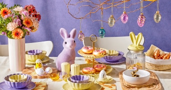 Paastafel vol met kleurrijke borden, kommetjes, paastakken met hangers, vaas met bloemen.