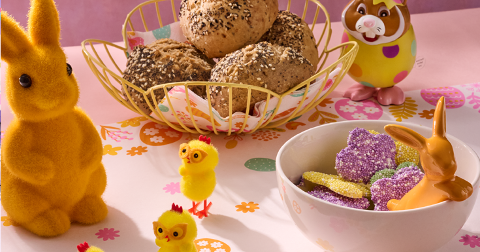 Paasontbijt tafel met een mandje vol meergranenbroodjes, kleurrijke paashaas- en kuikenfiguren en een kom met paaseitjes en paassnoepjes op een roze ondergrond.