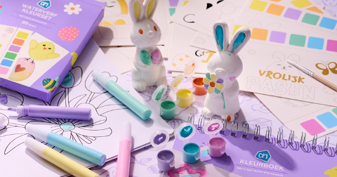 Paas knutselset van Albert Heijn met een aquarelverf kleurdos, kleurboek en decoratieve paashaasfiguurtjes, omringd door vrolijk gekleurde stiften, verfpotjes en tekenvellen.