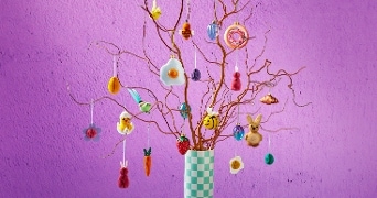 Decoratieve takken versierd met kleurrijke paashangers en papieren eieren in een geblokte vaas.